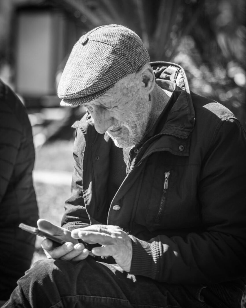A senior adult engrossed in using a smartphone while seated outdoors, captured in black and white.