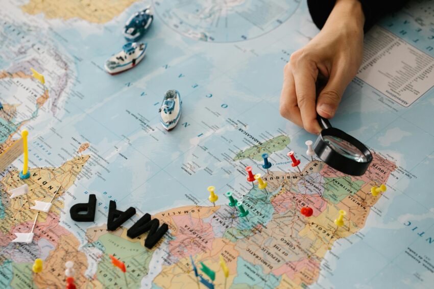 A hand using a magnifying glass over a world map with pins and toy ships.
