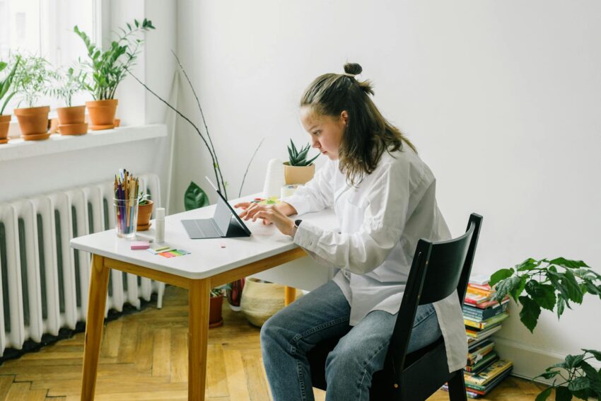 Teenage girl studying using tablet while sitting at a desk in a bright home setting.
