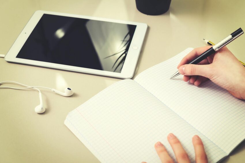 A person writing in a notebook beside a tablet and earphones, indoors.