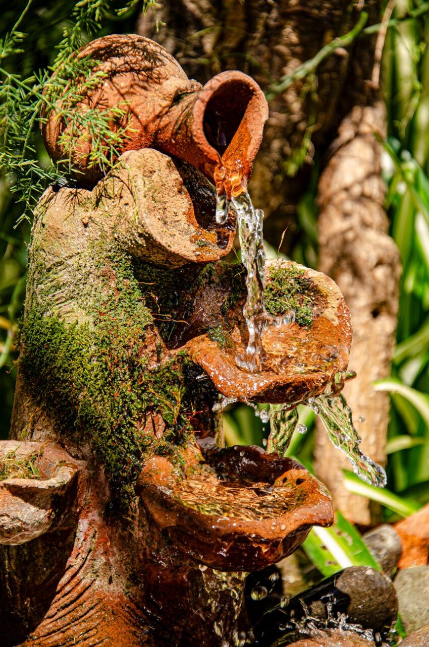 A close-up of a rustic terracotta fountain with flowing water, surrounded by lush greenery.