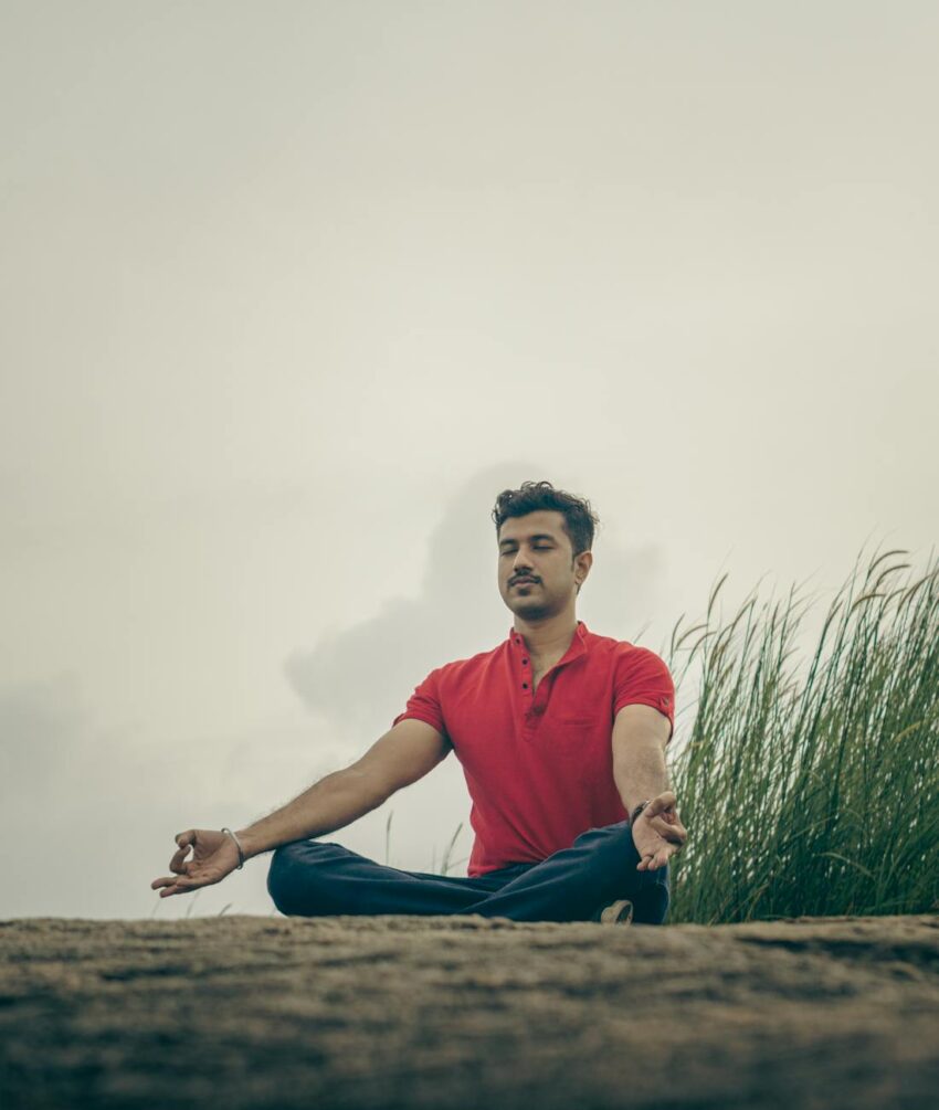 Adult man in a red shirt meditating outdoors on a grassy field, promoting relaxation and mindfulness.