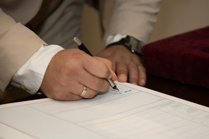 A close-up of a man signing a document, showcasing a wedding ring and pen.