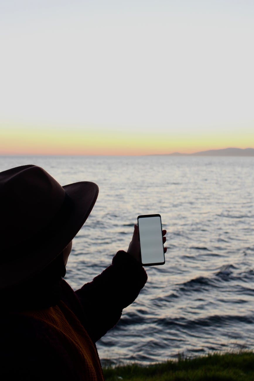 Silhouette of a person capturing a serene ocean sunset with a smartphone.