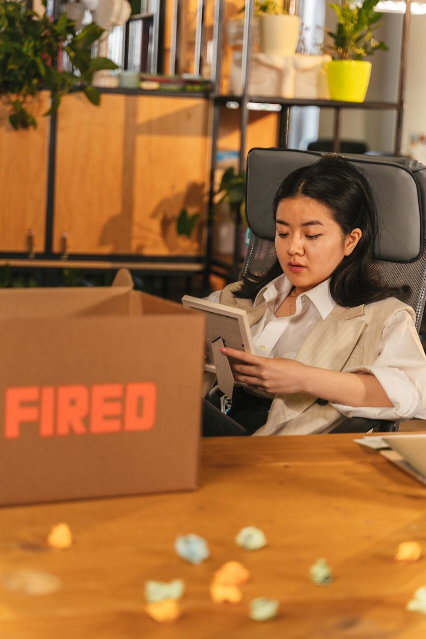 Woman sitting in office holding a picture frame next to a box labeled 'Fired'.