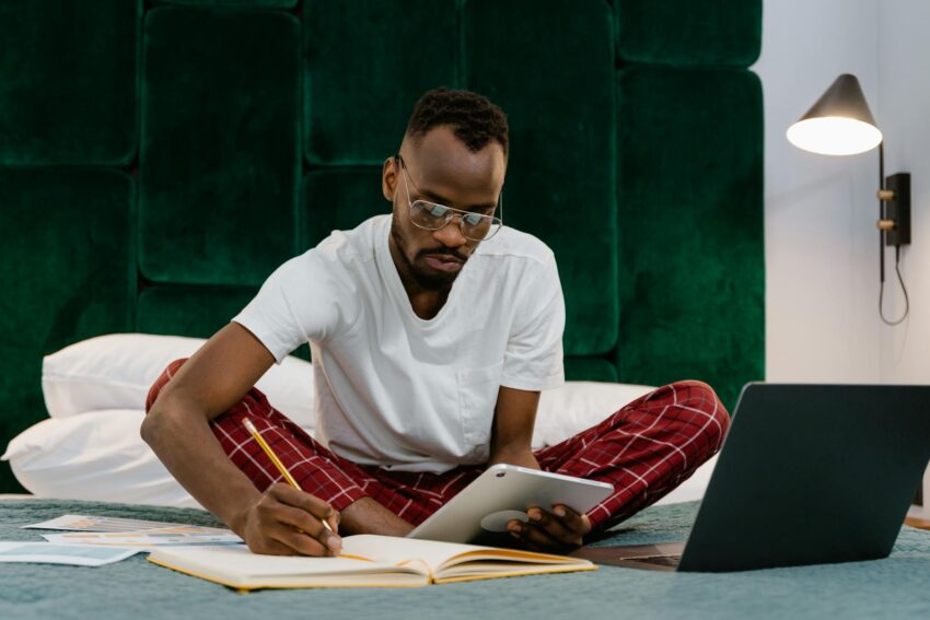A man working from home on a bed with a laptop and tablet, focused and engaged in remote work.