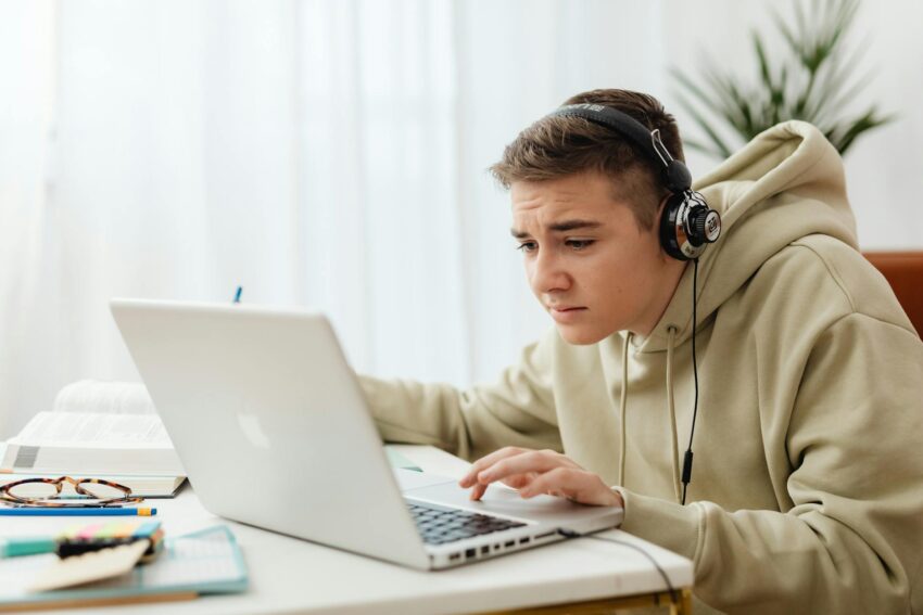Teen wearing headphones studies online with a laptop in a cozy indoor setting.