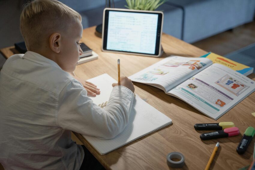 Young boy studying with notebook, pencil, and tablet on a wooden desk, focusing on education.