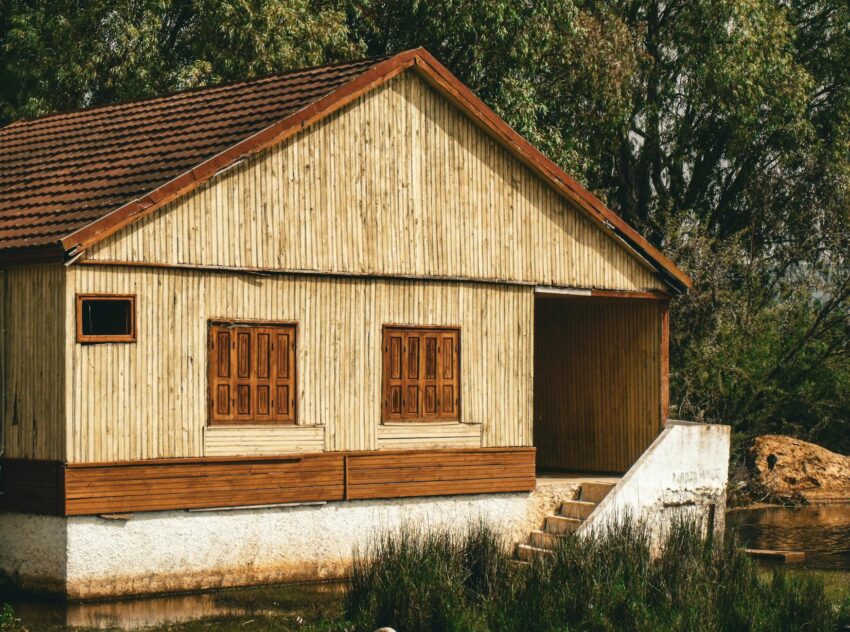 A rustic wooden house with red roof near a water body surrounded by greenery.