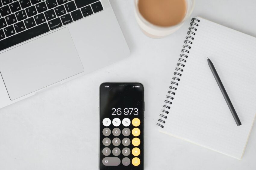 Overhead view of a laptop, smartphone displaying calculator, notebook, pen, and coffee on a white table.