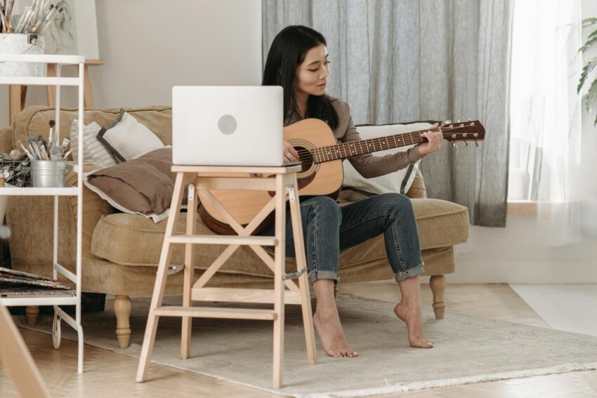 Woman practicing acoustic guitar in her living room via online lessons during the day.