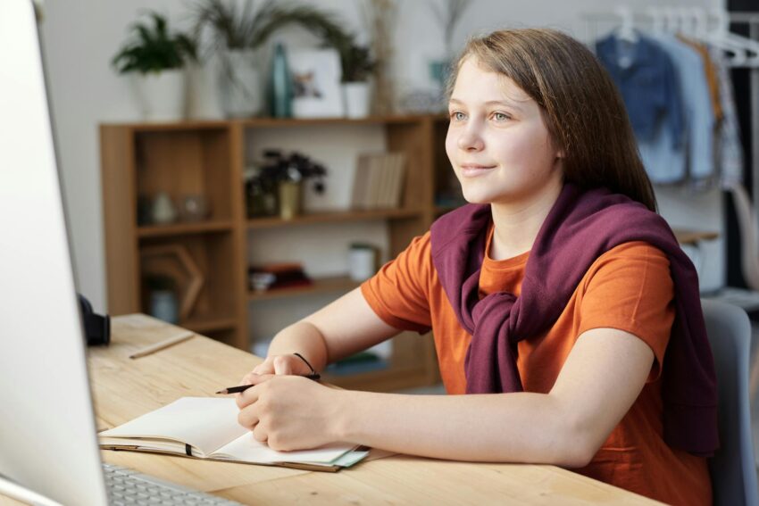 Teenage girl sitting at a desk with a laptop, studying and taking notes at home.