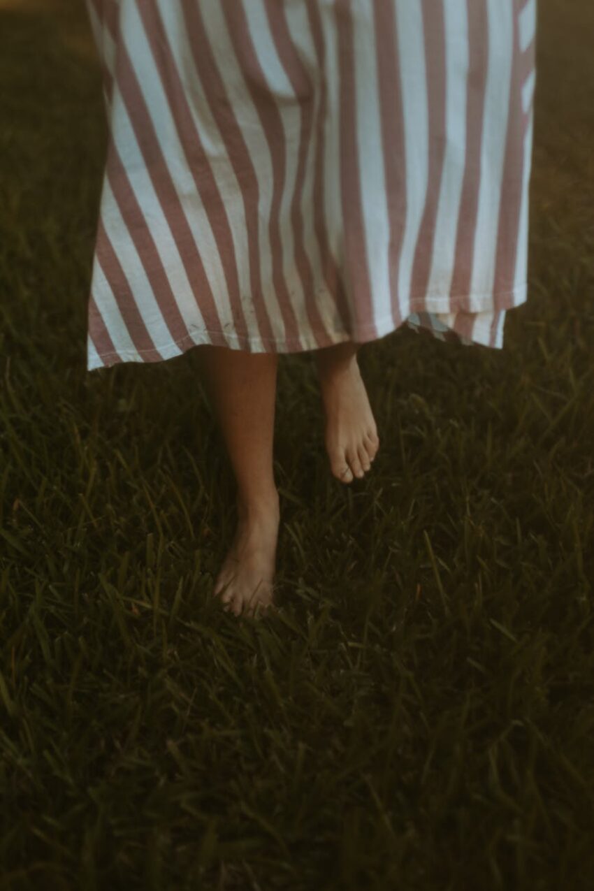 A person walks barefoot on lush grass, wearing a striped dress.