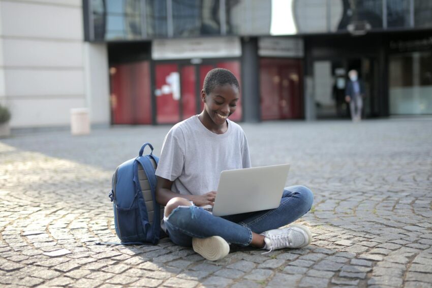 Smiling woman sitting on cobblestone street with laptop, engaging in online learning outdoors.
