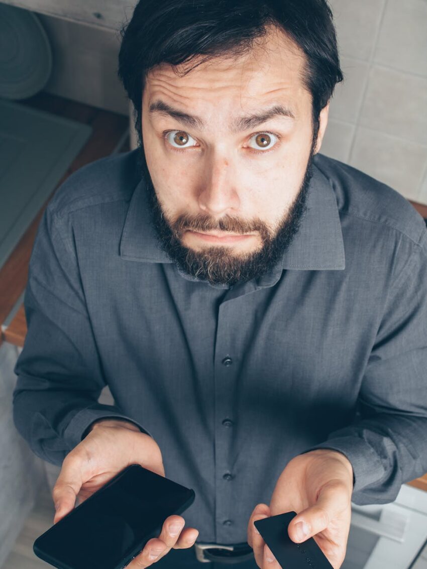 Bearded man in gray shirt looking confused while holding a smartphone and credit card.
