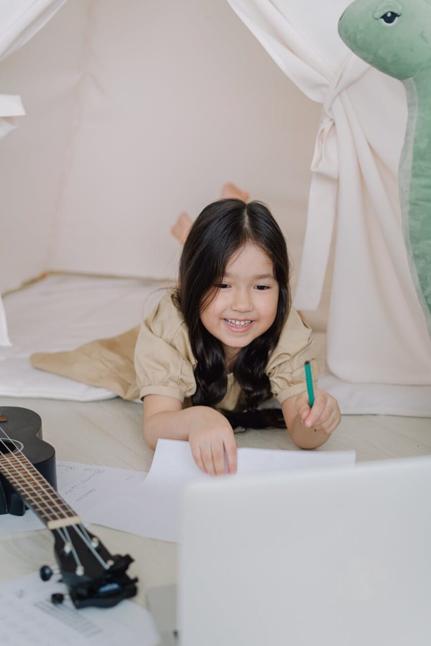 A cheerful girl learning at home, using a laptop and ukulele, expressing creativity and joy.