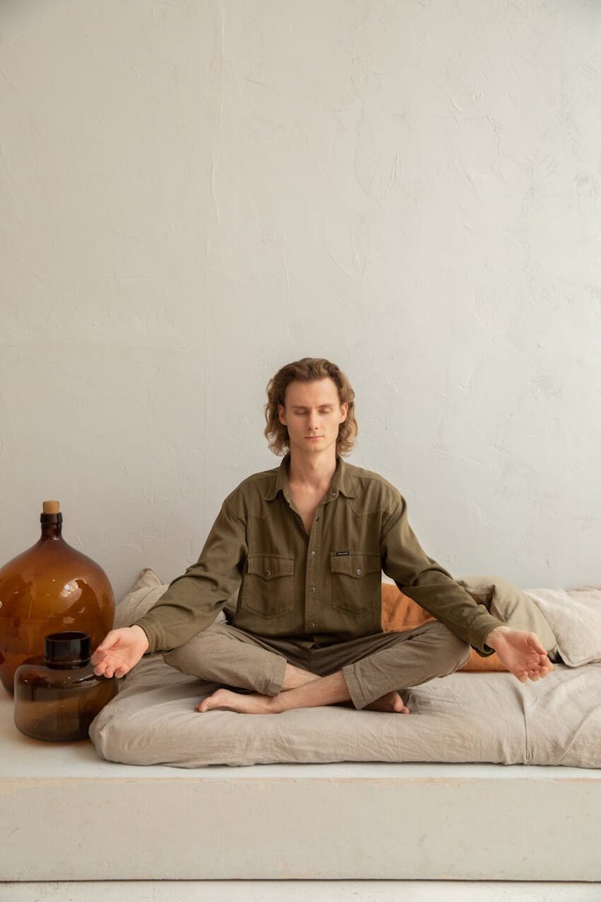 Young reflective barefoot male sitting in Padmasana pose with closed eyes on cozy mattress in house