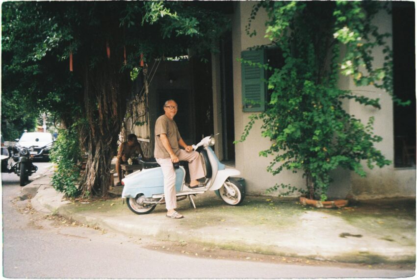 Elderly man sits on a classic scooter under a tree in a quaint urban setting.