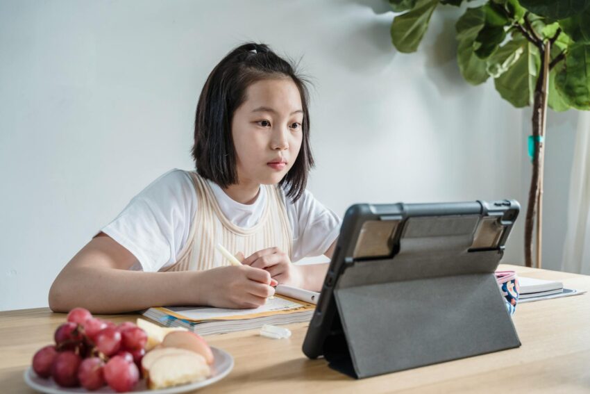 Young girl engaged in online learning using a tablet, seated at a table with snacks indoors.