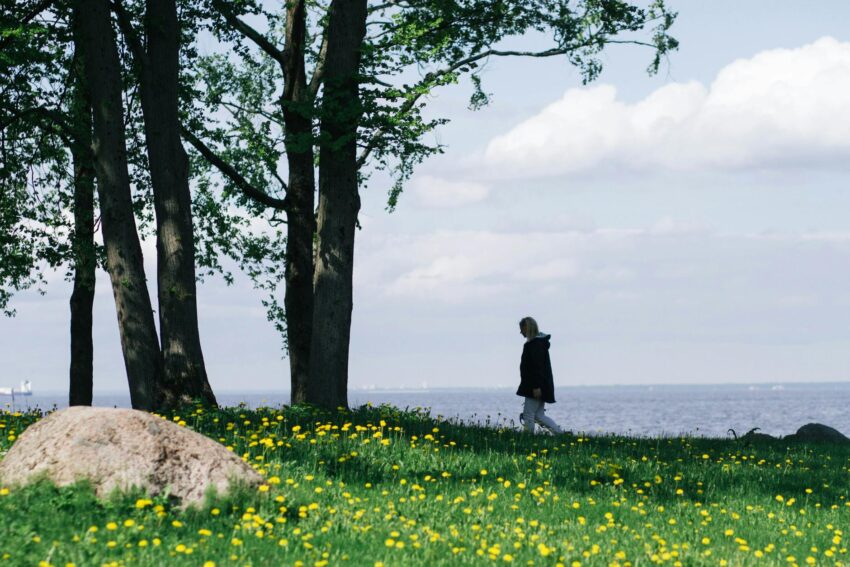 A woman walks along a grassy field by a serene lake under a clear spring sky.