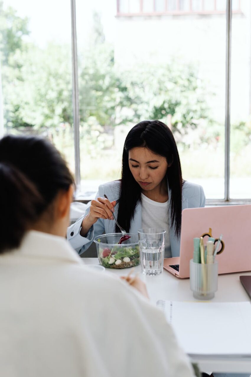 A young woman sits at her desk, enjoying a healthy salad lunch beside her laptop.