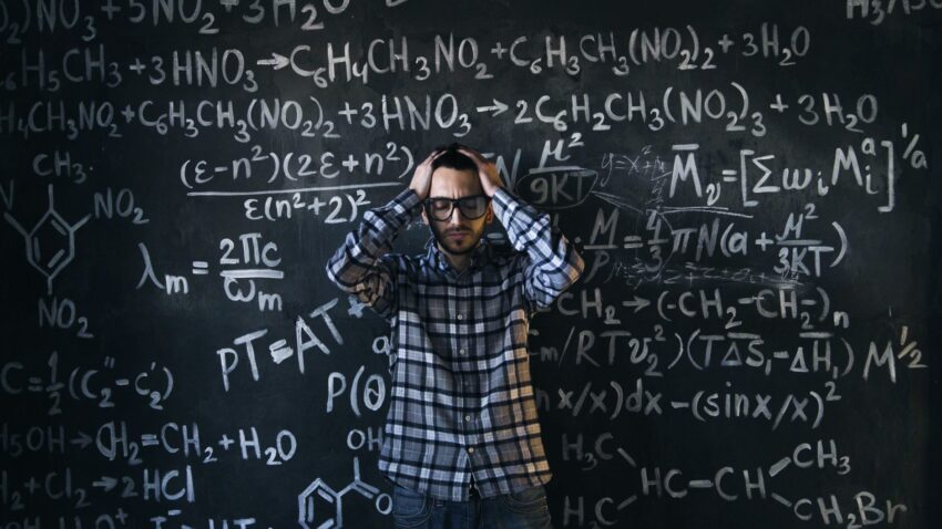 Man in glasses focusing on math problems, struggling with complex equations on chalkboard.