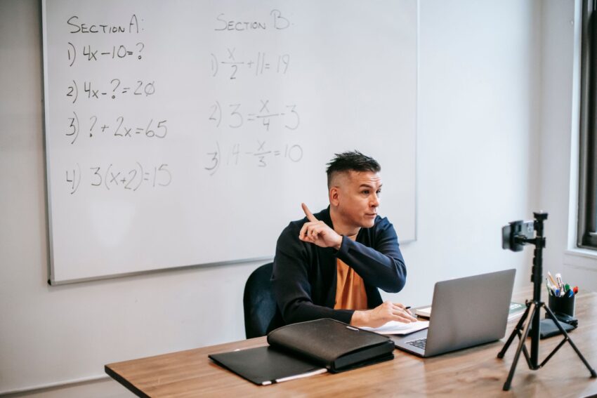 A male teacher explains math equations during an online class, using a laptop and whiteboard.