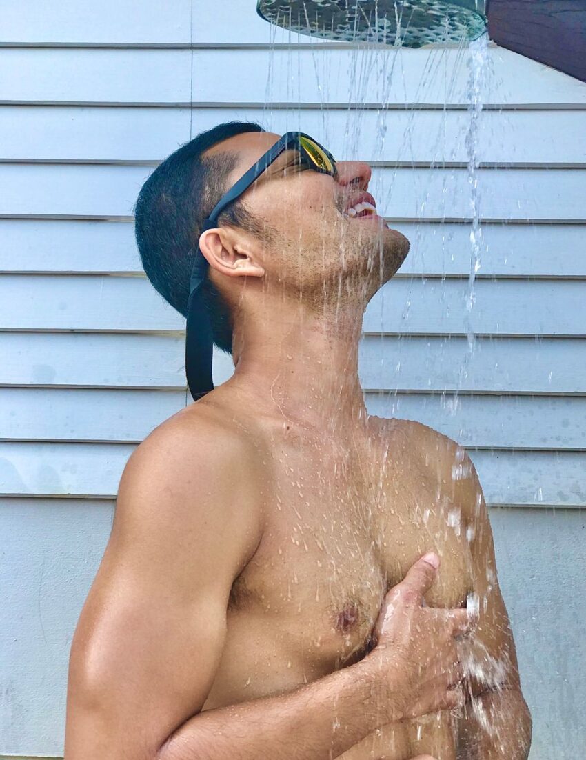 A person wearing sunglasses enjoys a refreshing outdoor shower on a sunny day.